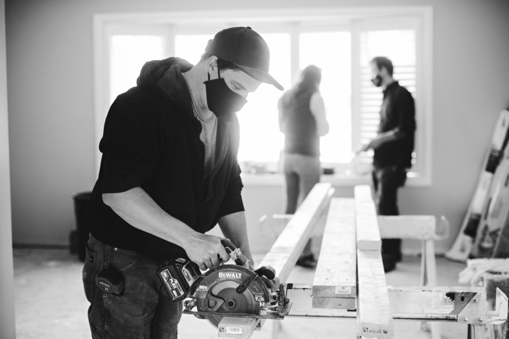 Carpenter cutting wood, contractor speaking to client in background. 